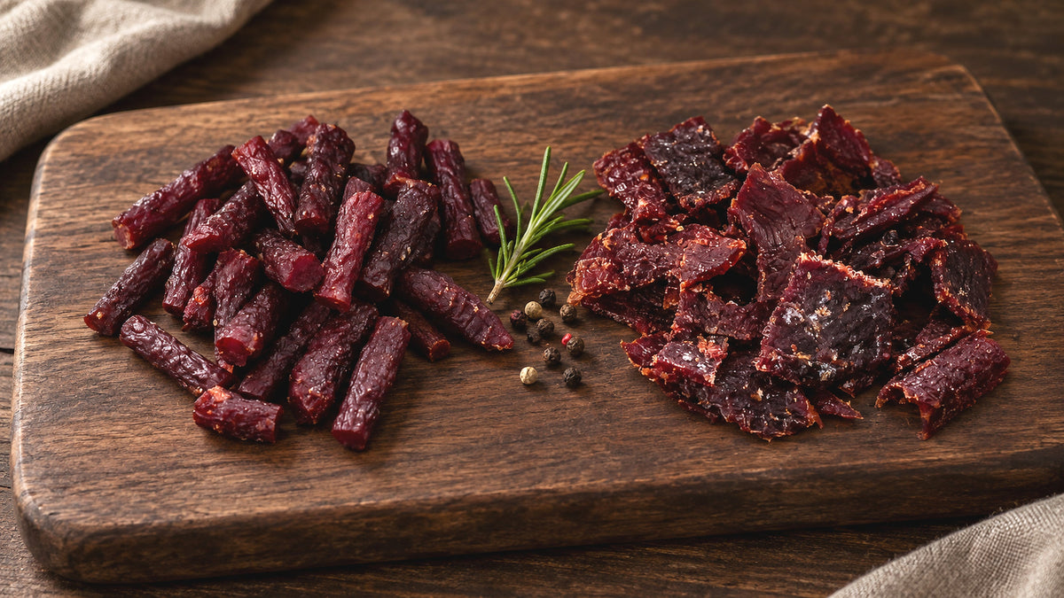 Beef meat sticks next to beef jerky pieces on a wooden board with black pepper